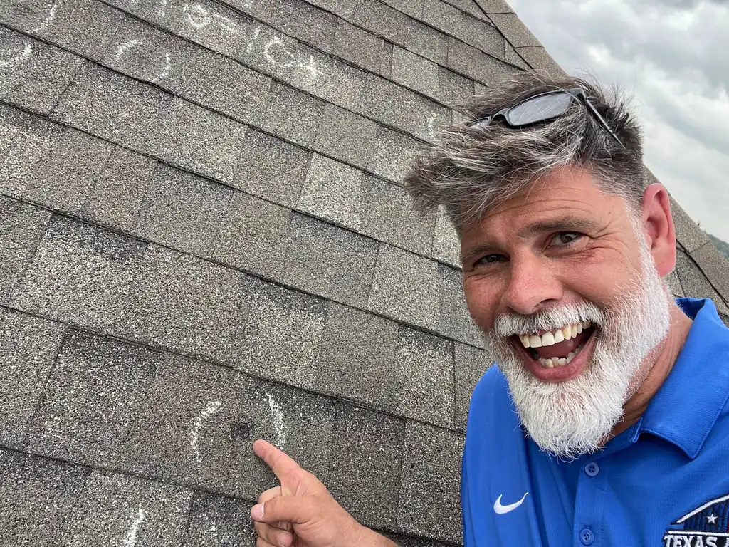 A Texas Accent Roofing inspector in a branded polo on an intact shingle roof, pointing at a hail bruise he has ringed in chalk beside a chalked ten-by-ten test square